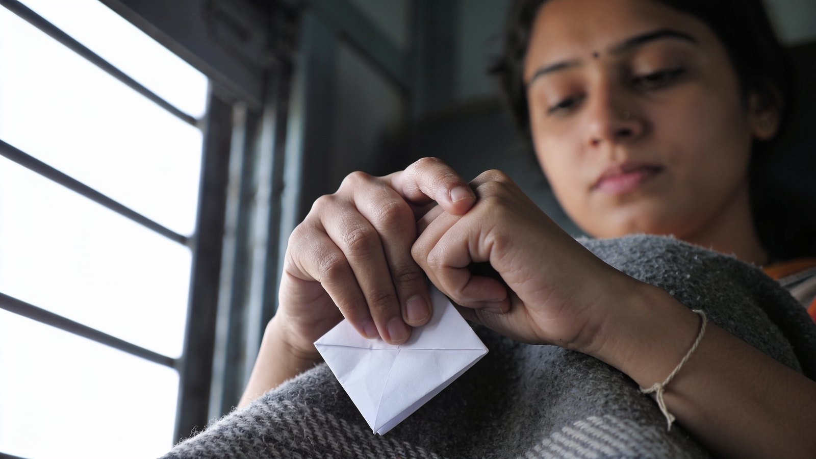 Close-up of a woman's hands tightly clutching a small folded blank slip of paper in her lap.