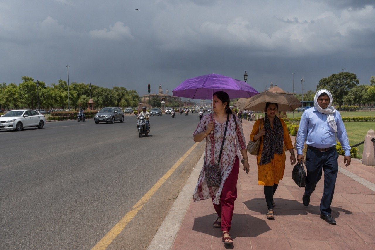 Residents under a hazy, heat-baked Delhi sky with India Gate in the distance, scattered clouds gathering on the horizon as the city awaits monsoon rain