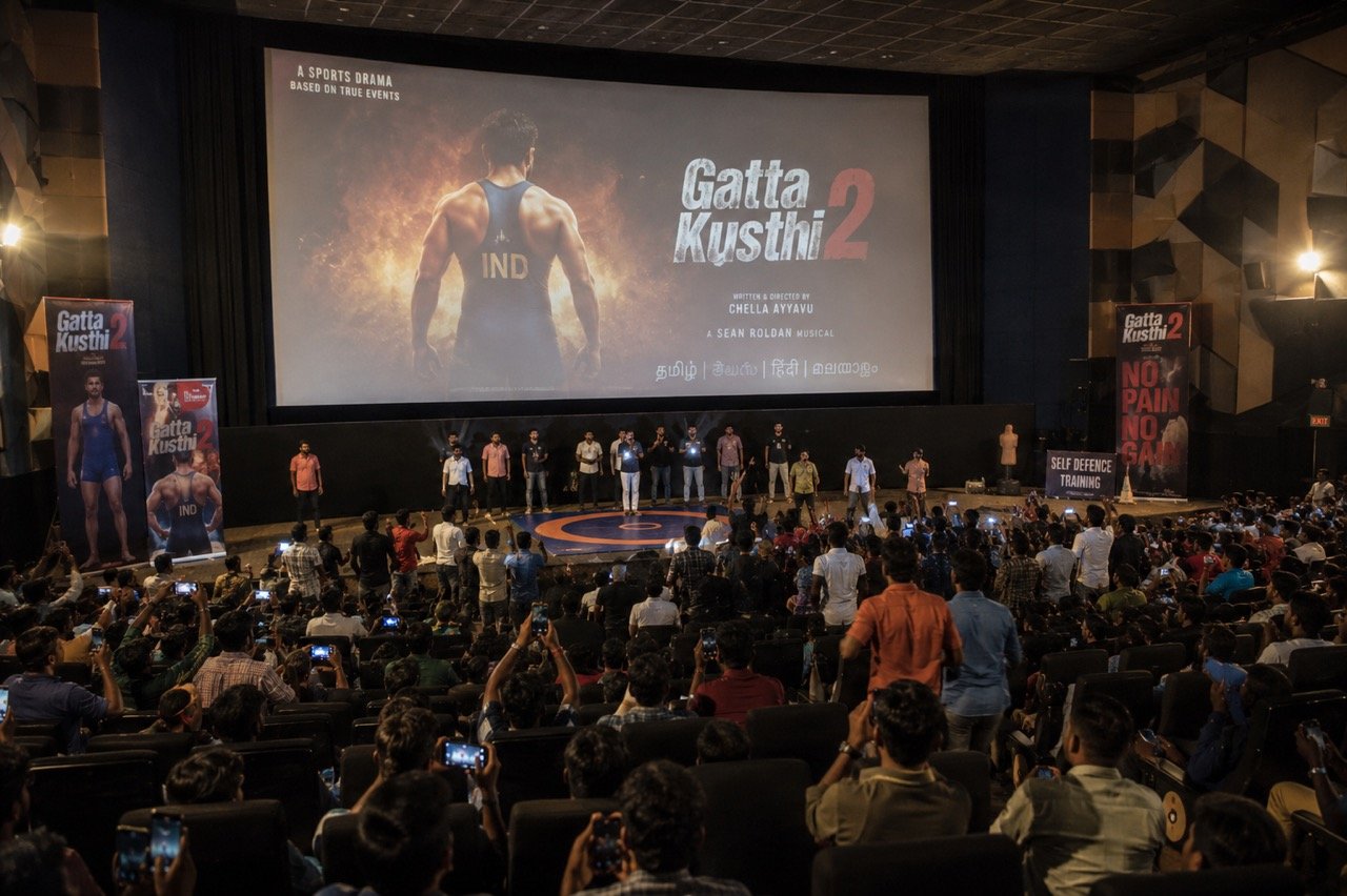 Two wrestlers grappling on a mud akhara under stadium lights, capturing the energetic sports-drama spirit of Gatta Kusthi 2