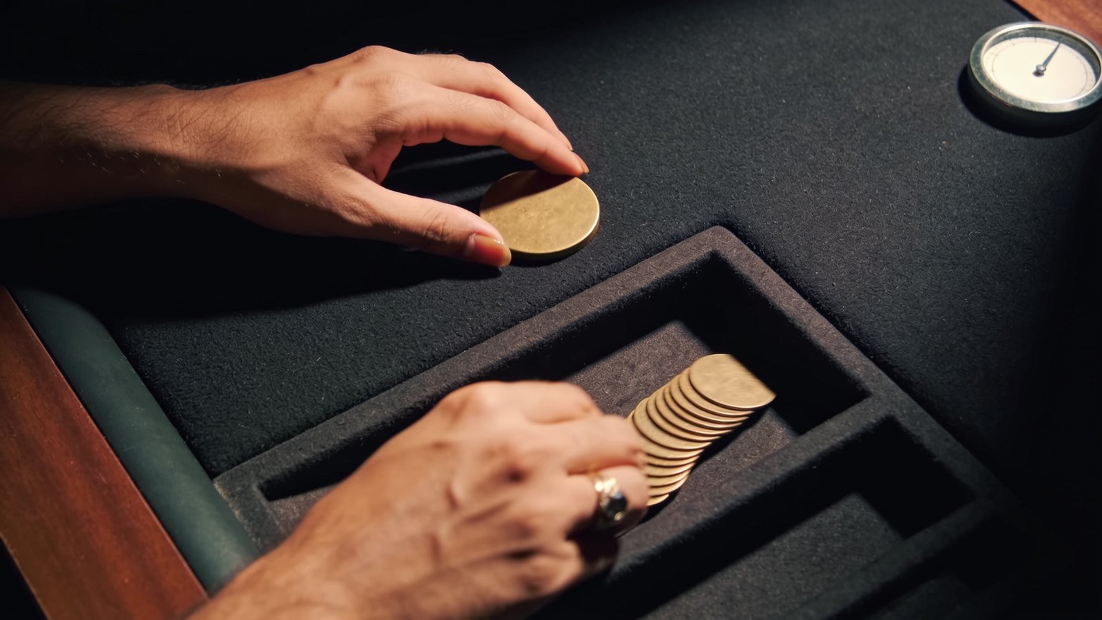 Close-up of hands moving plain brass tokens across a black felt table under a spotlight.
