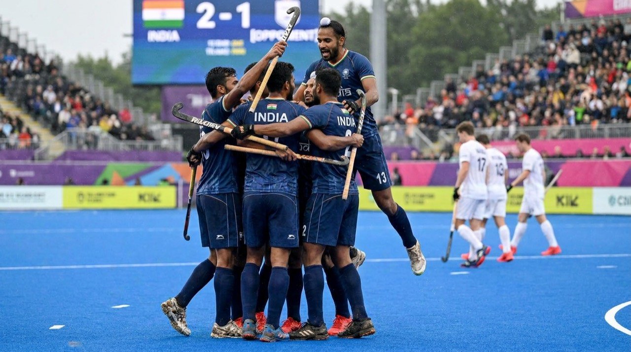 Field hockey players in blue celebrating a goal during an international match on a blue turf.