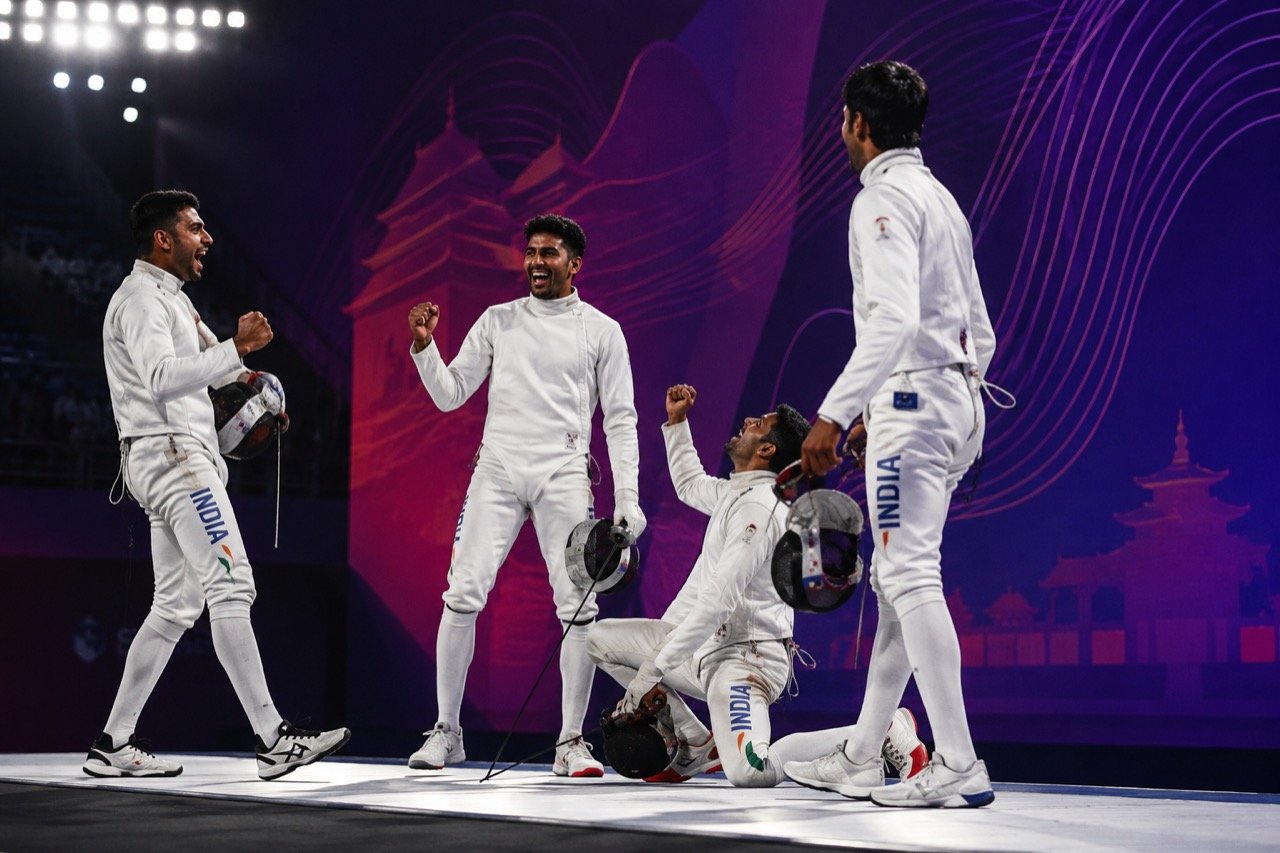 Two fencers in white protective gear duelling with foils on a competition piste under arena lights, with an Indian flag visible in the background