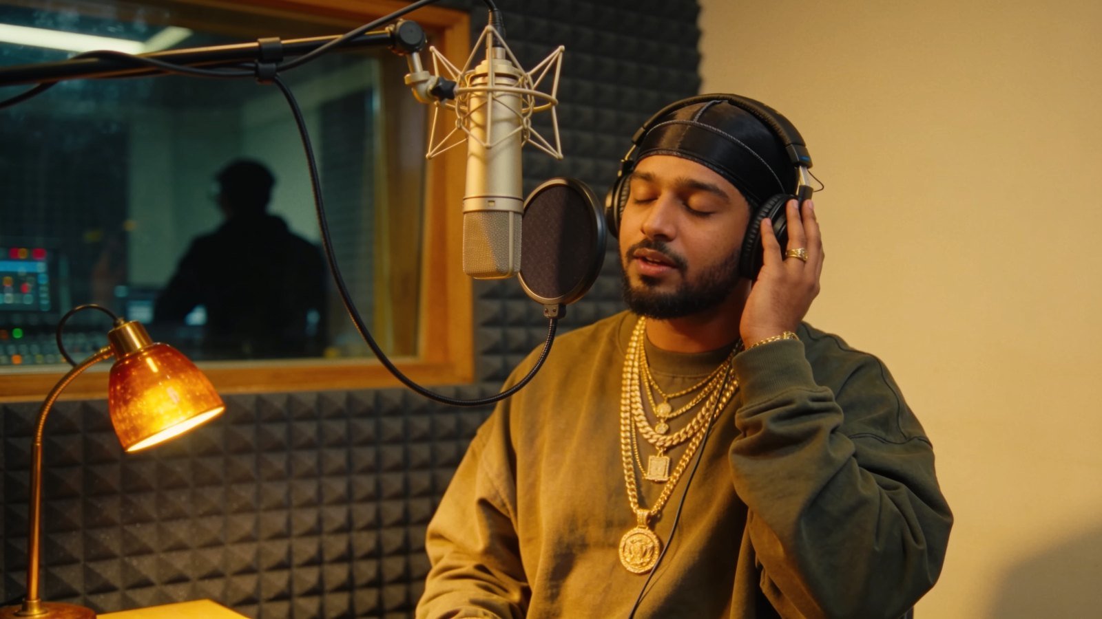 A young rapper in streetwear and headphones performs at a studio microphone inside a foam-lined vocal booth.
