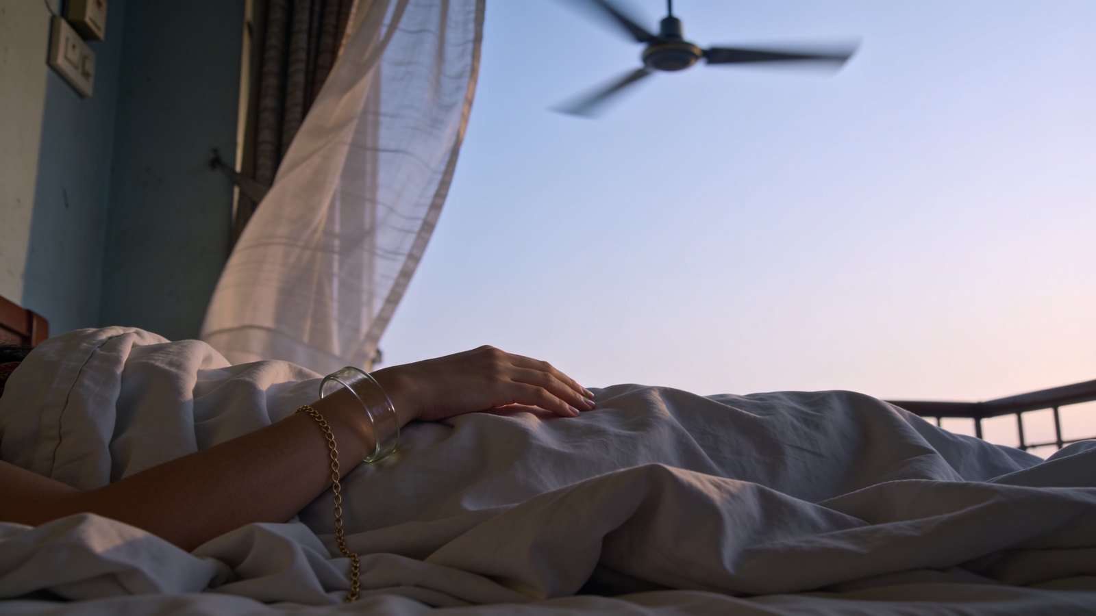 A woman's bangled hand resting on rumpled bedsheets at dawn, with a sheer curtain lifting nearby.