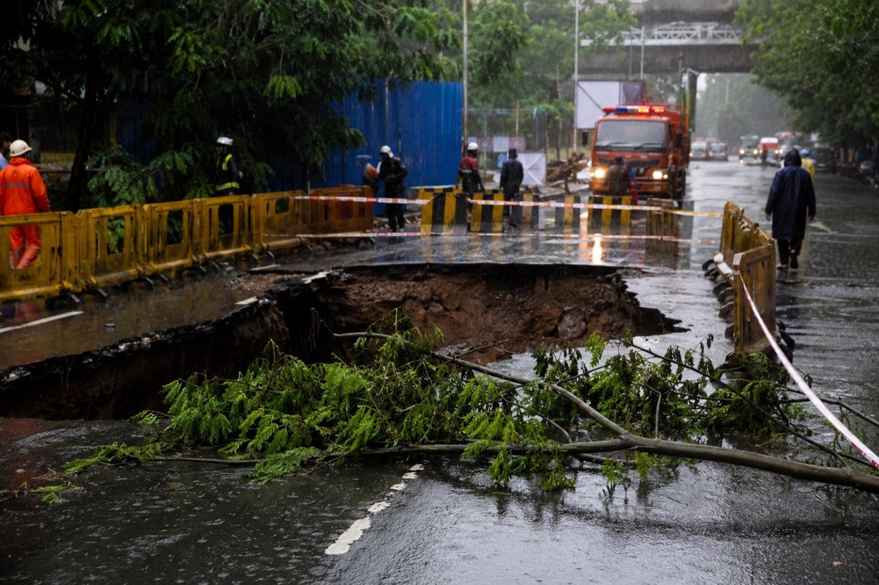 A collapsed section of a rain-soaked Mumbai arterial road with a vehicle tipped into an excavation pit, uprooted trees and warning barricades under grey monsoon skies