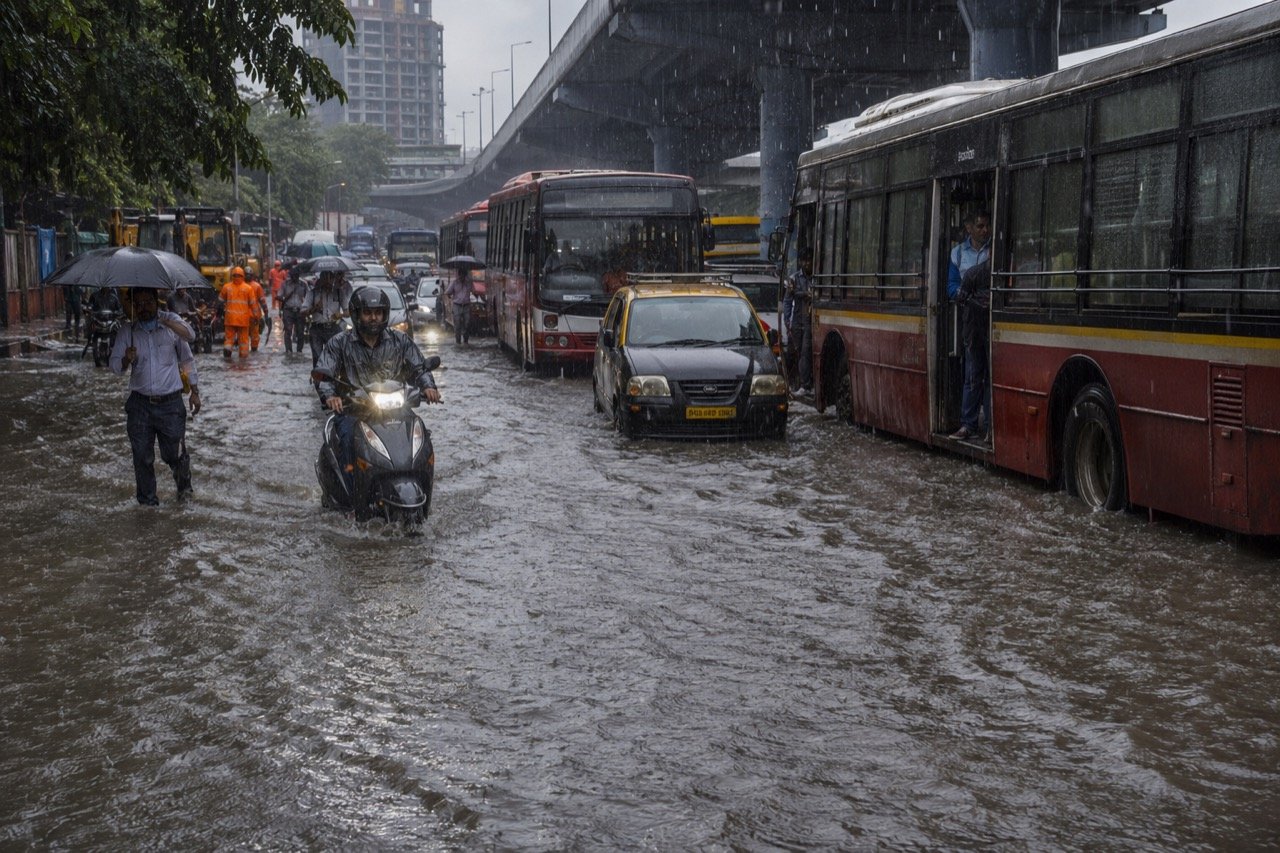 Commuters wading through a waterlogged Mumbai suburban street in heavy monsoon rain, with stalled traffic, umbrellas and a local train line in the background