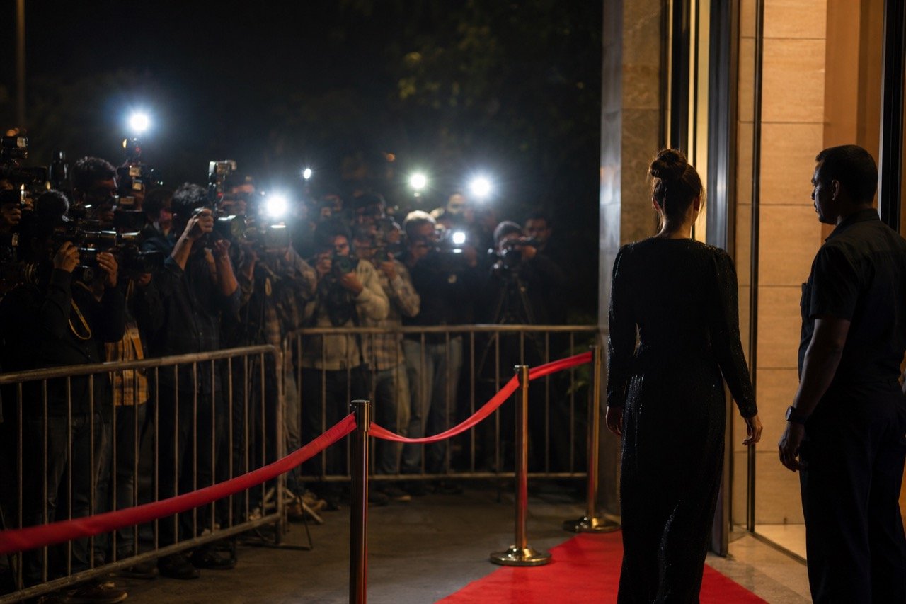 A crowd of paparazzi photographers with raised cameras and flashing lights outside a Mumbai event venue at night
