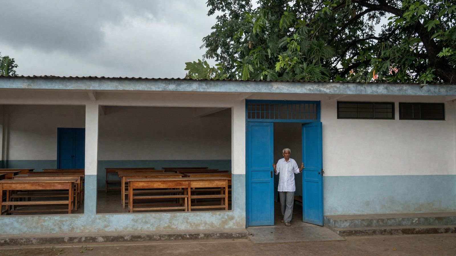 Caretaker unlocking the gate of a Delhi government school examination hall with rows of empty desks inside under monsoon light.