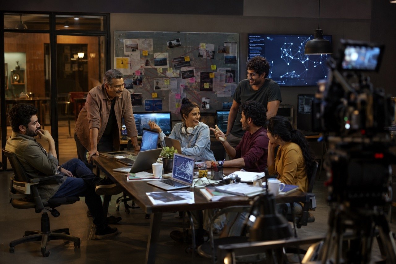 Two mismatched investigators silhouetted against glowing computer screens in a dim office, capturing the cybercrime comedy tone of Pritam and Pedro.
