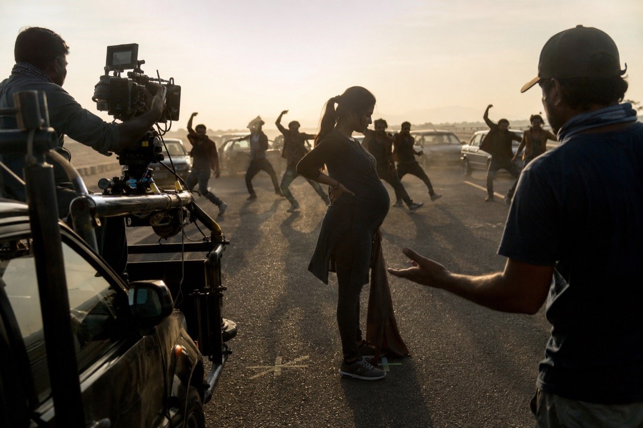 A film set at dusk with camera rigs, crew silhouettes and a car staged for an action sequence, illustrating the physical demands of a South Indian movie shoot
