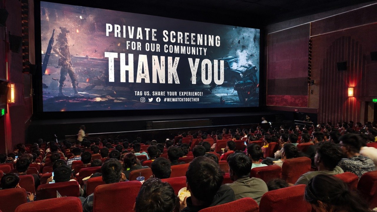 A packed single-screen cinema hall glowing under a projector beam as an excited young crowd watches a Bollywood spy thriller on the big screen